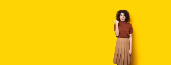 Schockierte kaukasische Frau mit lockigem Haar, die auf den gelben Freiraum in ihrer Nähe zeigt — Stockfoto