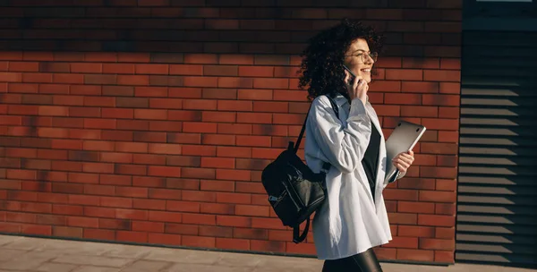 Junge kaukasische Studentin mit lockigem Haar telefoniert beim Gehen mit Tasche und Laptop — Stockfoto