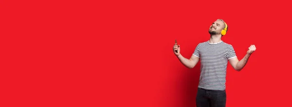 Homem caucasiano com barba e cabelo loiro está gesticulando uma vitória enquanto ouve música em uma parede vermelha com espaço livre — Fotografia de Stock