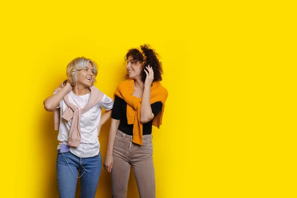 Encantadoras mujeres caucásicas con el pelo rizado posan en una pared amarilla con espacio en blanco mientras escuchan música a través de auriculares — Foto de Stock