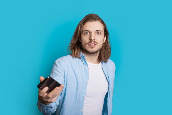 Homem caucasiano de cabelos longos com cerdas está olhando seriamente para a câmera enquanto segura um celular em um fundo azul — Fotografia de Stock