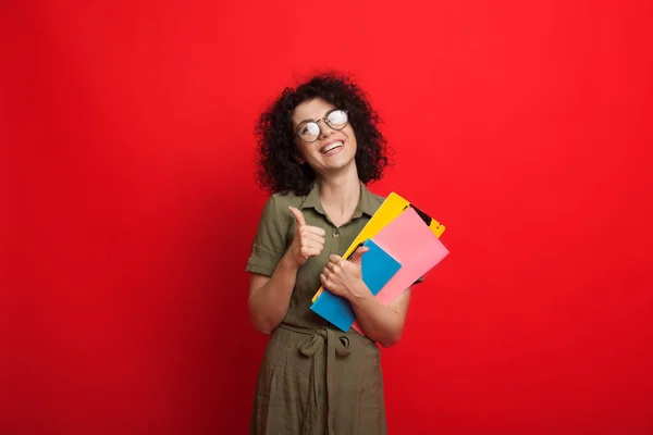 Lockige kaukasische Studentin hält ein paar Bücher in der Hand und gestikuliert mit einem ähnlichen Zeichen, während sie auf rotem Hintergrund posiert — Stockfoto