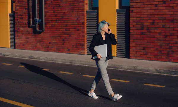 Blauw harige blanke student lopen in de straat, terwijl het hebben van een telefoon discussie en het houden van een laptop — Stockfoto