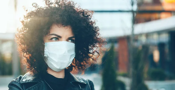 Kıvırcık saçlı beyaz kadın dışarıda binanın önünde poz verirken grip maskesini takıyor. — Stok fotoğraf