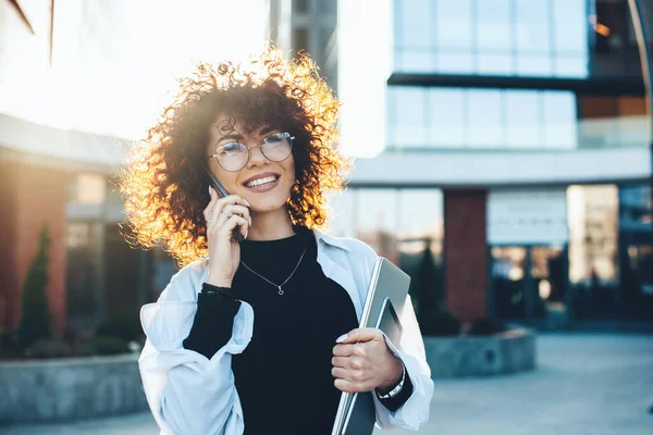 Cabello rizado mujer de negocios caucásica tener una discusión telefónica mientras posando con gafas y aparatos modernos en frente de un edificio — Foto de Stock