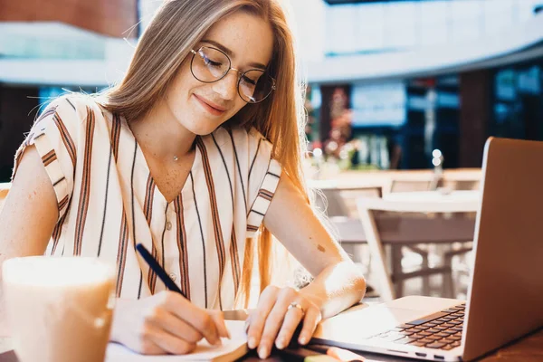 Ginger καυκάσιος φοιτητής με φακίδες κρατώντας σημειώσεις, ενώ εργάζονται με έναν υπολογιστή σε ένα εστιατόριο — Φωτογραφία Αρχείου