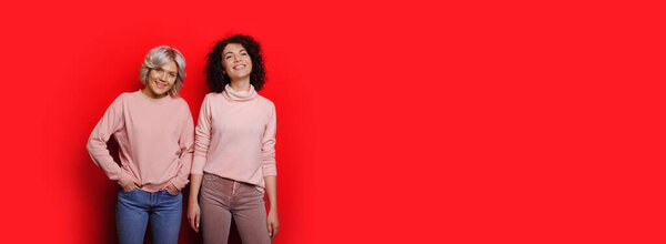 Two sweet caucasian women posing on a red background with free space in the same sweaters