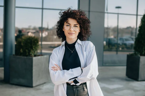 Affascinante donna caucasica con i capelli ricci e gli occhiali è in posa con le mani incrociate e guardando la fotocamera — Foto Stock