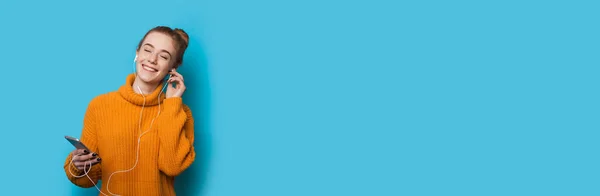 Mulher caucasiana ruiva com sardas ouvindo música usando telefone e fones de ouvido está posando em uma parede azul com espaço em branco — Fotografia de Stock