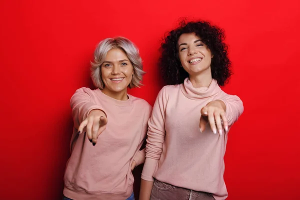 Lockige brünette Frau und ihr blonder Freund zeigen auf die Kamera, während sie auf rotem Hintergrund posieren — Stockfoto