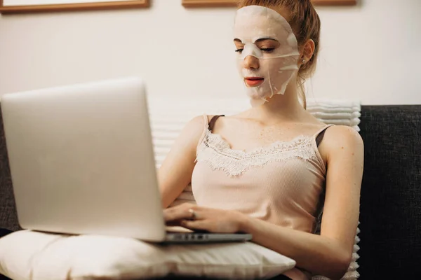 Blank meisje met gember haar dragen van een hydratatie masker terwijl chatten met iemand aan de laptop in bed — Stockfoto