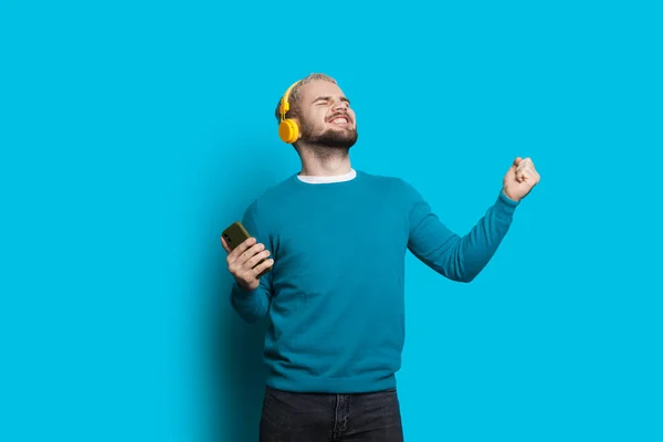 Homem caucasiano emocional com barba e cabelo loiro está ouvindo música em uma parede azul usando celulares e fones de ouvido — Fotografia de Stock