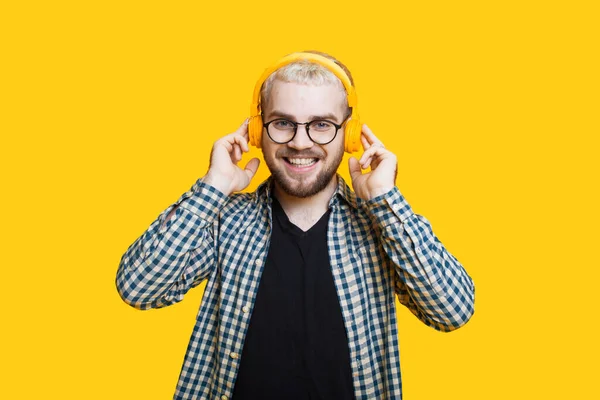 Close up foto de um homem caucasiano barbudo com cabelo loiro usando fones de ouvido e óculos posando em uma parede amarela — Fotografia de Stock