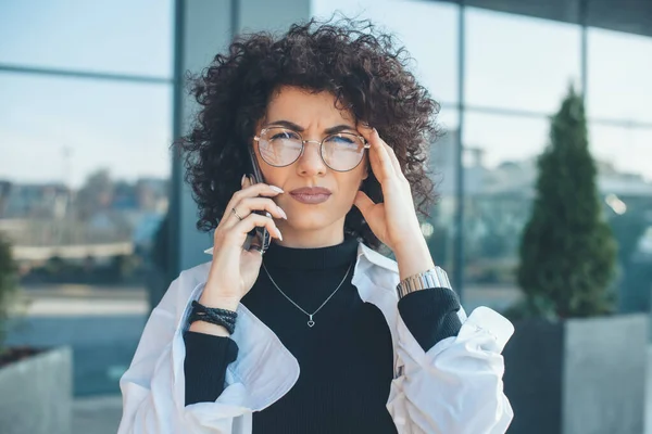 Grave donna caucasica con i capelli ricci e gli occhiali guardando la fotocamera mentre ha una discussione di telefono aziendale al di fuori — Foto Stock