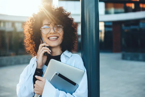 Glada kaukasiska kvinna med lockigt hår har en telefon diskussion medan poserar med en bärbar dator och tablett utanför — Stockfoto