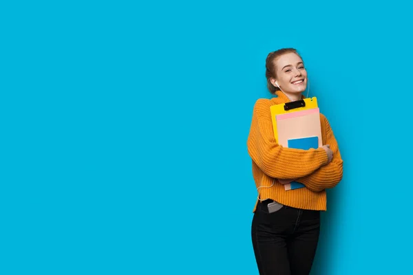 Härlig kaukasisk student med rött hår och fräknar poserar med hörlurar och några böcker på en blå vägg med fritt utrymme — Stockfoto