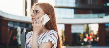 Gözlüklü ve çilli bir kadın güneşli bir günde telefon ve dizüstü bilgisayarla poz veriyor.