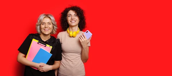 Charmiga kaukasiska studenter med lockigt hår poserar på en röd vägg med tomt utrymme håller en mobil och några böcker — Stockfoto