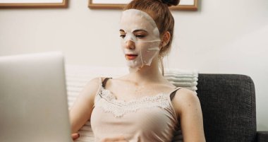 Bilgisayarın başında otururken kırışıklık önleyici maske takan kızıl saçlı beyaz bir kadın.