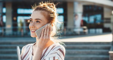Kafkasyalı iş kadını, çilli ve kızıl saçlı dışarıda yürürken telefonla konuşuyor.