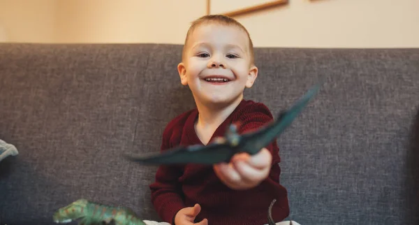 Caucásico niño pequeño sentado en el sofá y jugando con los dinosaurios juguetes —  Fotos de Stock