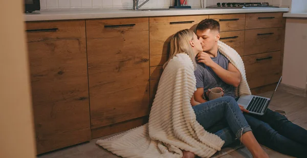 Covered caucasian couple kissing on the floor and drinking a tea while using a computer in the kitchen — Stock Photo, Image