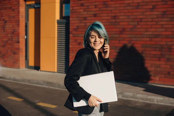 Härligt blåhårig kaukasisk entreprenör talar på telefon och promenader med en bärbar dator — Stockfoto