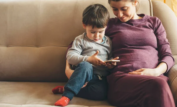 怀孕的高加索女人，带着雀斑和她的小男孩坐在沙发上，用电话 — 图库照片