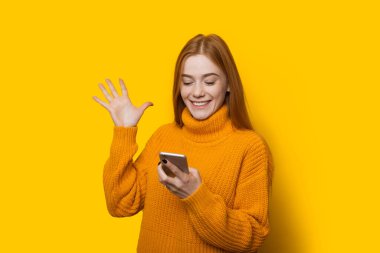 Kafkasyalı çilli kız cep telefonuyla zafer işareti yapıyor ve sarı arka planda kalın bir kazakla poz veriyor.