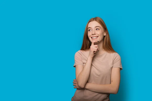 Piegowata biała kobieta z rudymi włosami myśląca o czymś i uśmiechająca się na niebieskiej ścianie z wolną przestrzenią — Zdjęcie stockowe