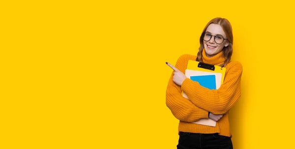 Estudiante de jengibre con pecas y anteojos apuntando al espacio libre amarillo cerca de ella con el dedo abrazando algunos libros — Foto de Stock