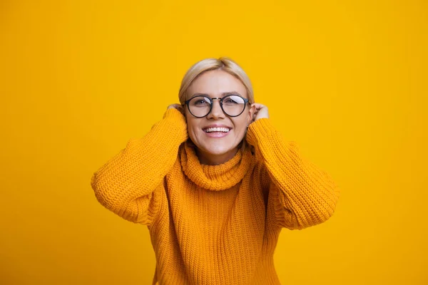 Zbliżenie się zdjęcie białej kobiety z blond włosami i okularami dotykającej jej włosów i uśmiechającej się do kamery na żółtym tle — Zdjęcie stockowe
