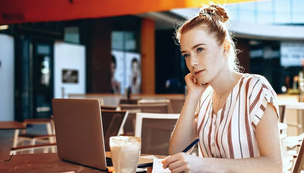 Κοκκινομάλλα καυκάσια επιχειρηματίας με φακίδες γράφει και σκέφτεται κάτι σε μια καφετέρια, ενώ πίνοντας ένα κοκτέιλ και χρησιμοποιώντας ένα φορητό υπολογιστή — Φωτογραφία Αρχείου