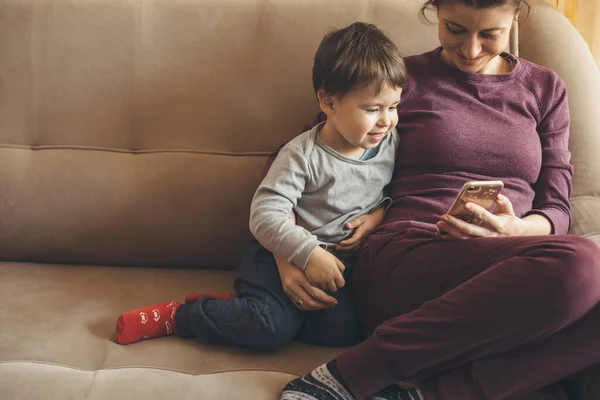 Ibu Kaukasia dan anak kecilnya berbaring di sofa dan menggunakan ponsel selama karantina — Stok Foto