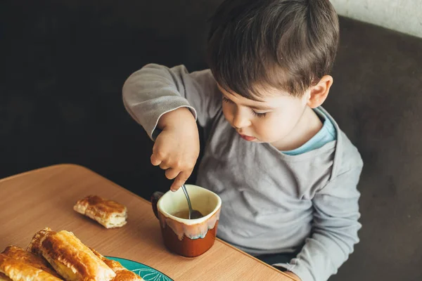 Foto vista superior de um menino caucasiano sentado à mesa e bebendo um chá com colher enquanto come alguns biscoitos — Fotografia de Stock