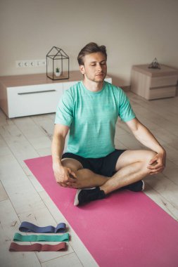 Sarışın, beyaz bir adam evde yoga yapıyor. Yerde pembe bir halının üzerinde. Yanında da lastik bantlar var.