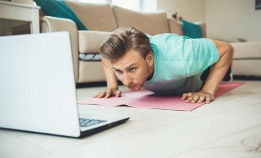Yerde dizüstü bilgisayara bakarken spor halısında şınav çeken beyaz bir adam.