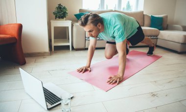 Yoga halısı üzerinde esneme egzersizleri yapan konsantre sarışın beyaz bir adam, yanında su bardağıyla dizüstü bilgisayara bakıyor.