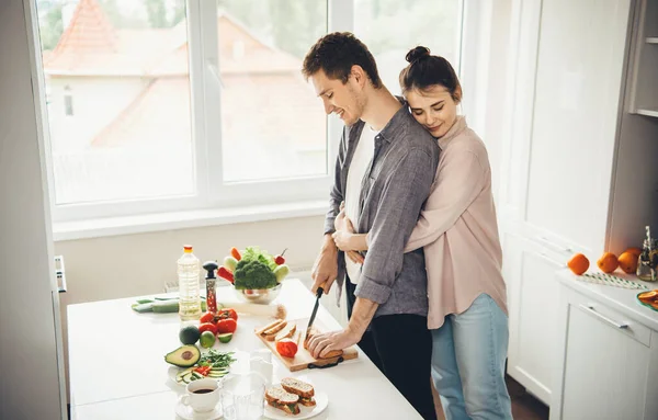 Bruna ragazza abbracciando il suo amante in cucina mentre fa panini — Foto Stock