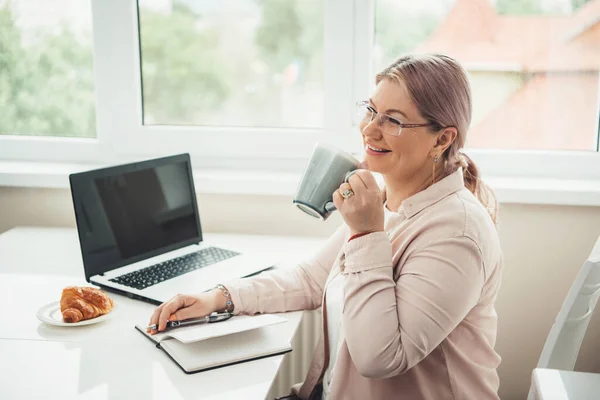 Caucasian senior woman working at the laptop from home while drinking tea with croissant and smile — Stock Photo, Image