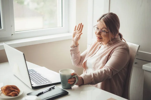 Caucasian senior woman greeting somebody on the laptop having an online meeting while drink a tea with croissant — Stock Photo, Image