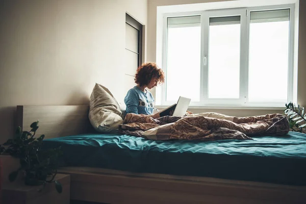 Mujer caucásica con el pelo rizado leyendo un libro en su cama sentado cerca de la ventana con una computadora — Foto de Stock