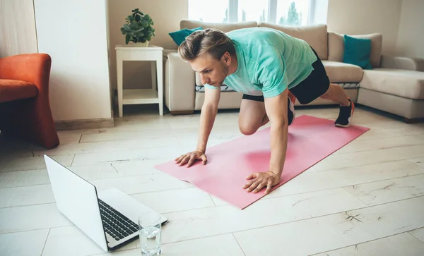 Concentrato uomo caucasico bionda facendo esercizi di stretching sul tappeto yoga mentre guardare il computer portatile con un bicchiere d'acqua nelle vicinanze — Foto Stock