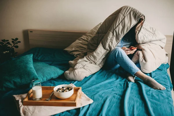 Mujer caucásica cubierta con una colcha está charlando por teléfono antes de comer leche con cereales en la cama —  Fotos de Stock