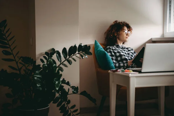 Ung affärskvinna med lockigt hår och glasögon arbetar hemifrån vid datorn tänker på något medan du håller en bok — Stockfoto