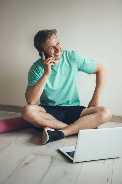 Uomo caucasico biondo che parla al telefono prima di fare esercizi di stretching sul pavimento con un computer portatile — Foto Stock