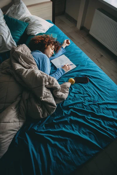 Mujer caucásica dormida con cabello rizado sintiéndose cansada después de hacer la tarea sosteniendo una pluma y descansando cerca de un libro y móvil — Foto de Stock