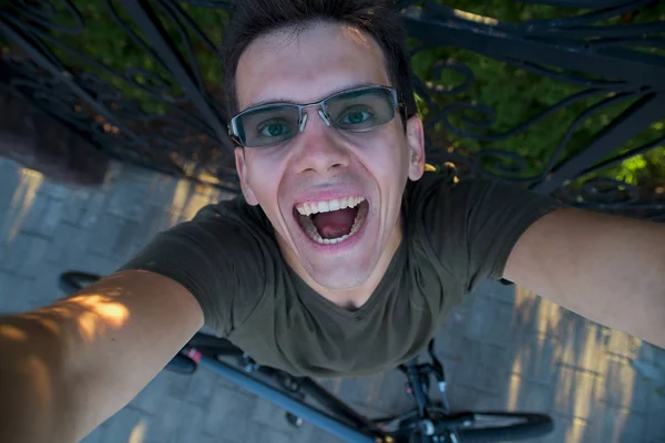 Smiling exited man in green shirt with sunglasses with bike bellow on green background of bushes and black metal fence stands with raised hands out of the frame.