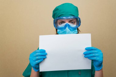 Yeşil takım elbiseli, mavi maskeli, koruyucu gözlüklü, sarı arka planda beyaz kağıtlı kadın doktor. Boşluğu kopyala.