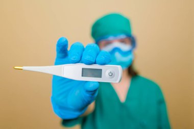 Yeşil takım elbiseli kadın doktor ve mavi maske eldivenleri ve sarı arka planda termometresi olan koruyucu gözlükler. Seçici odak.